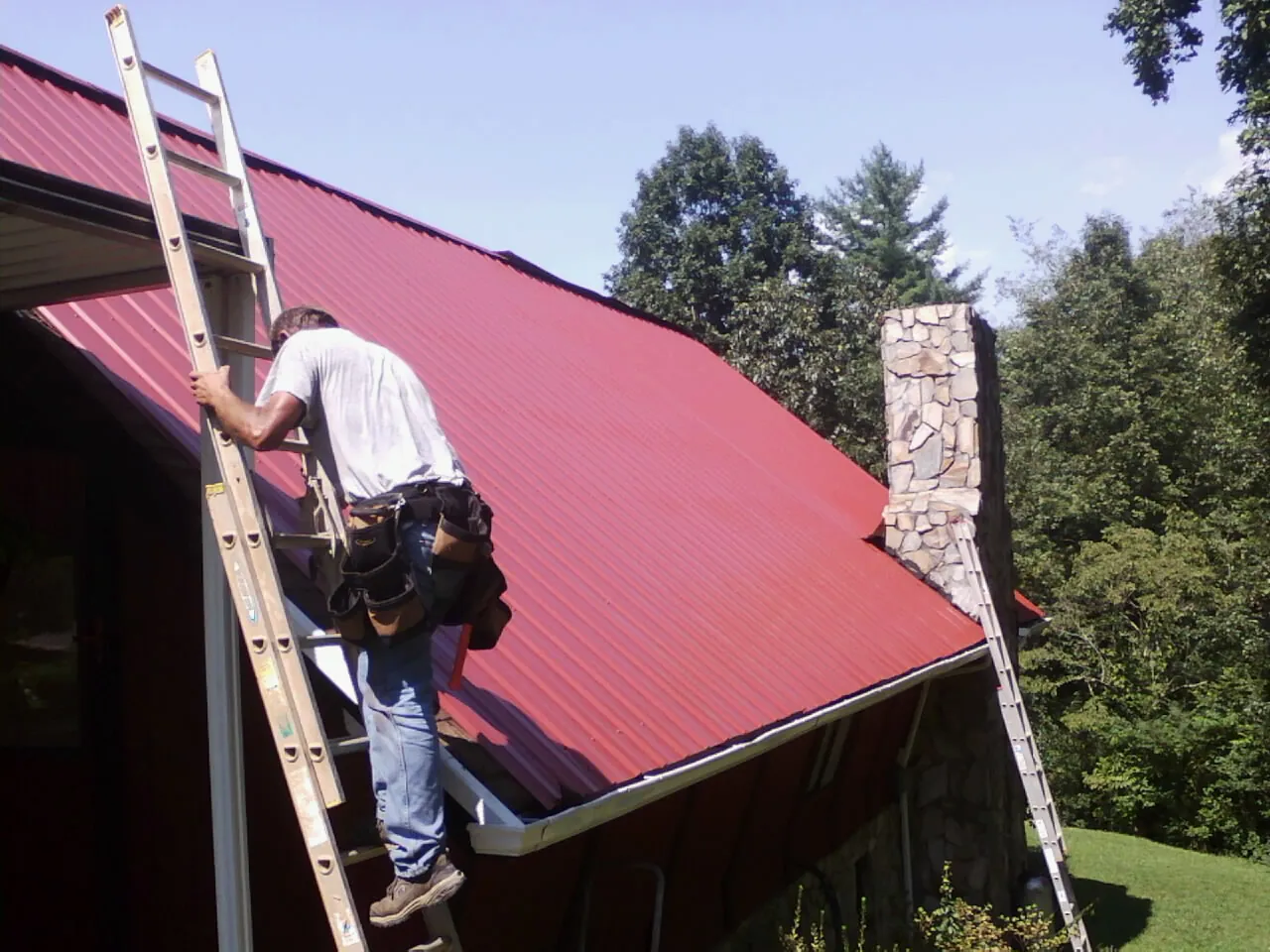 Roofer on a ladder beside a red metal roof and stone chimney — Soffit & Fascia Repair by Roofing Brothers in Commerce
